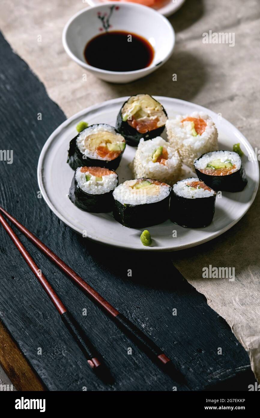 Hausgemachte Sushi-Brötchen mit Lachs, japanischem Omelett, Avacado, Wasabi und Sojasauce mit Holzstäbchen auf grauem Papier über schwarzem Holzbackgrou Stockfoto