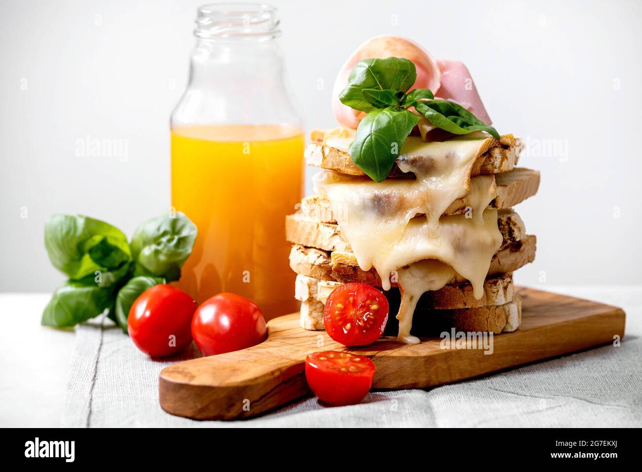 Vorrat an gerösteten, geschmolzenen, Käse gepressten Sandwiches mit Schinkenfleisch, Kirschtomaten, Orangensaft und Basilikumblättern auf Holzschneidebrett auf weißem m Stockfoto