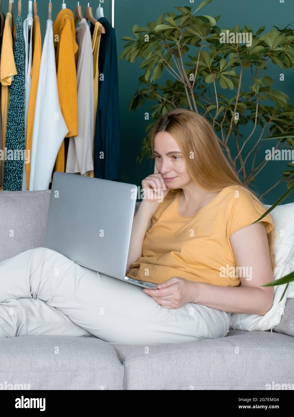 Blonde Frau mittleren Alters sitzt auf dem Sofa und kommuniziert online per Laptop. Stockfoto