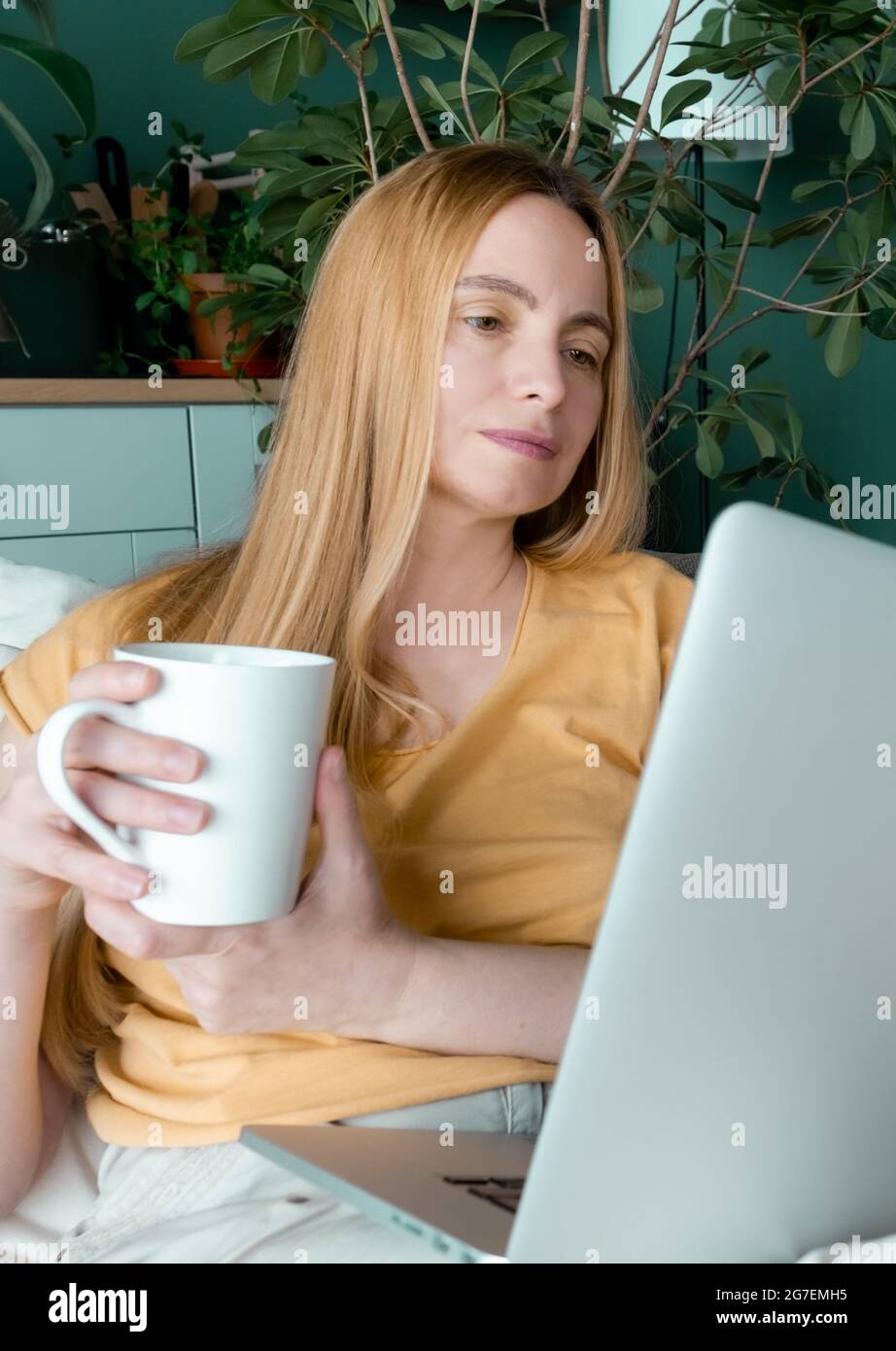 Blonde Frau mittleren Alters sitzt auf dem Sofa und kommuniziert online per Laptop. Stockfoto