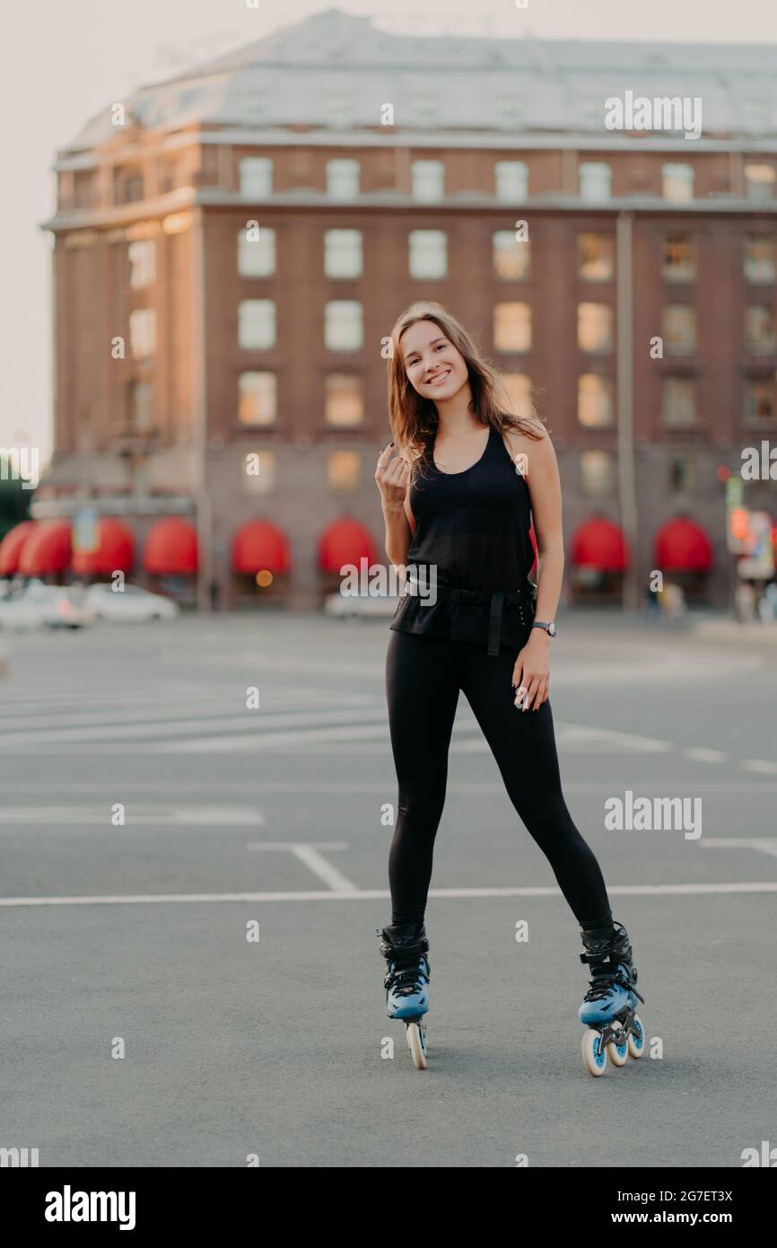 In voller Länge Aufnahme der aktiv lächelnden Frau genießt extreme Sportfahrten auf Rollerblades in schwarz gekleidet bequeme Kleidung steht im Freien lernt Stockfoto