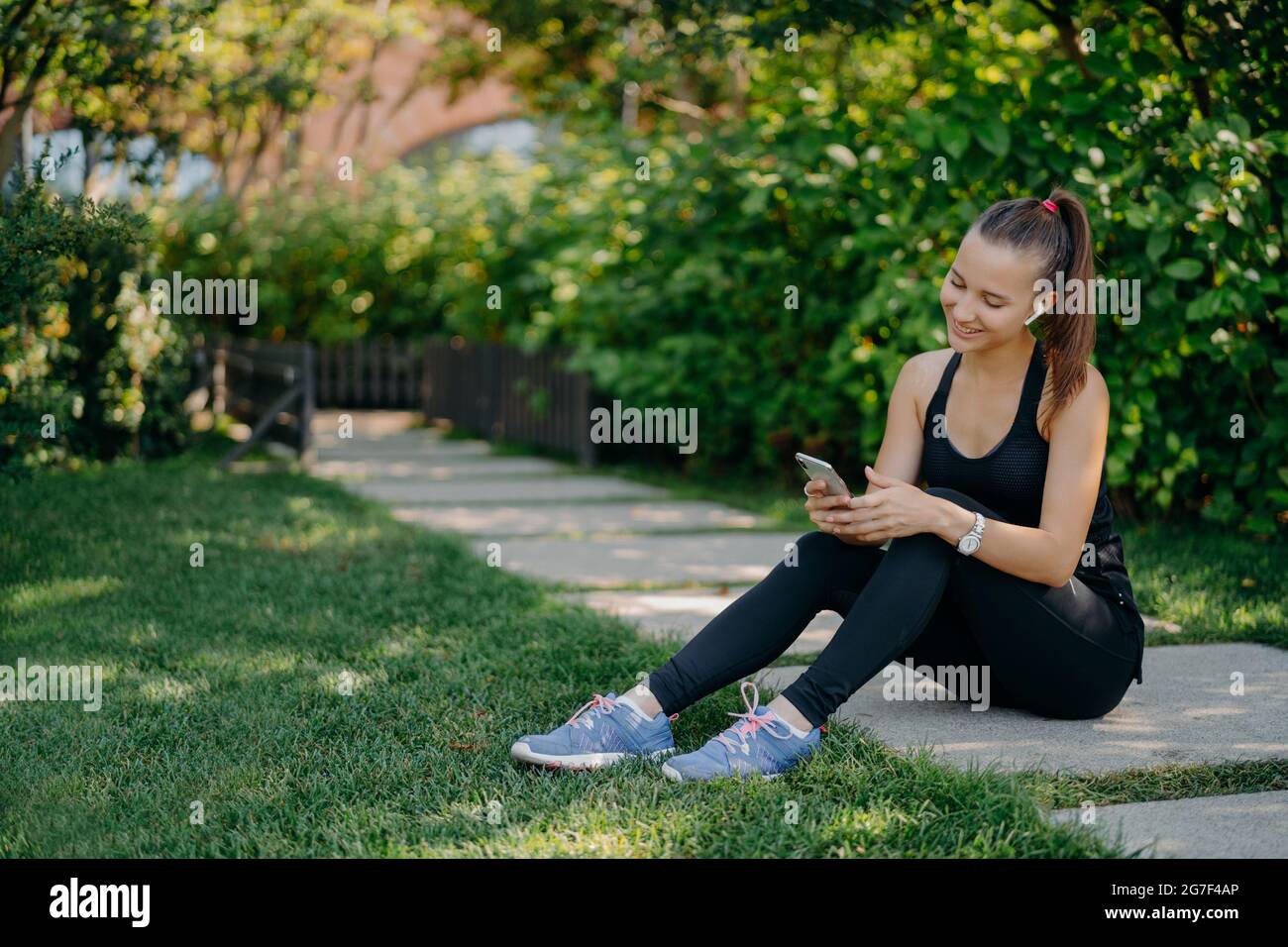 Erfreut athletische Frau in den Sportskloten lächelt fröhlich surft Mobiltelefon, während Musik über drahtlose Kopfhörer hört macht Pause nach der Sonne Stockfoto