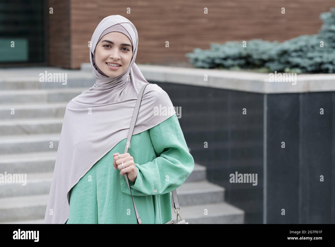 Porträt einer positiven, modernen, jungen islamischen Geschäftsfrau, die den Handgriff der Umhängetasche gegen das moderne Bürogebäude hält Stockfoto