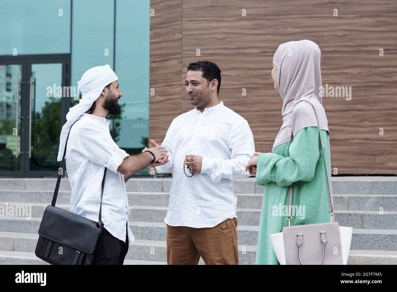 Zufriedene junge muslimische Geschäftspartner, die auf der Treppe gegen das Gebäude stehen und nach dem Treffen Handschlag geben Stockfoto