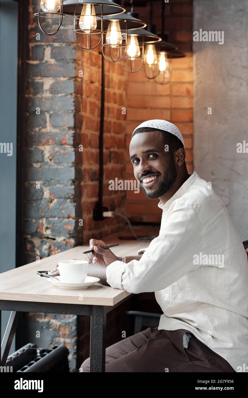 Porträt eines lächelnden, gut aussehenden bärtigen Schwarzen im kufi-Hut, der mit Stift am Tisch im Loft-Café sitzt Stockfoto