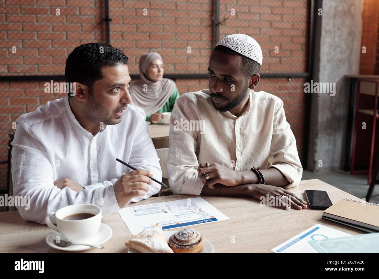 Beschäftigte junge, multiethnische Geschäftsleute in islamischen Outfits, die im Café am Tisch sitzen und über die Tafel diskutieren Stockfoto