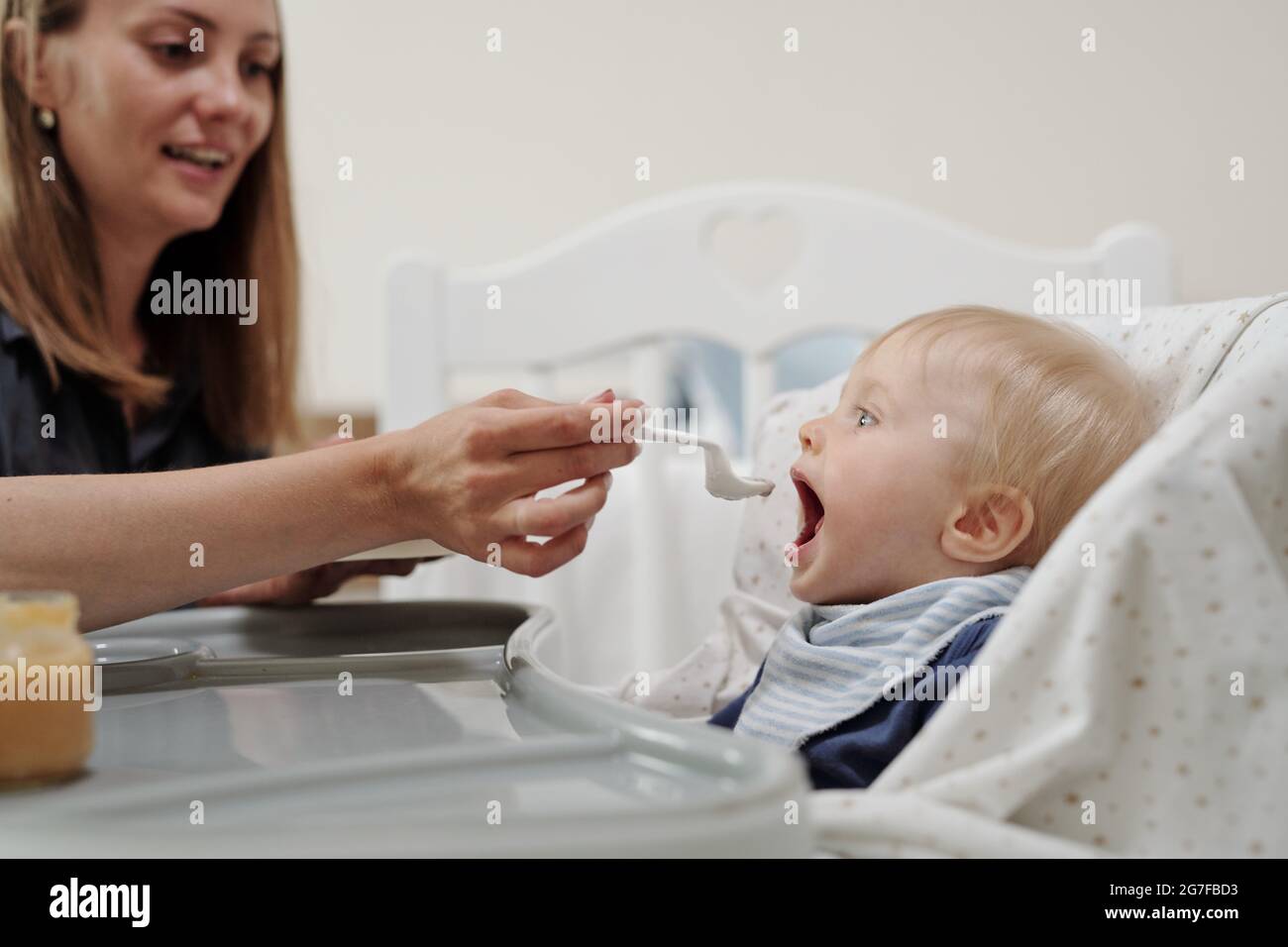 Positive junge Mutter, die dem Sohn einen Löffel Babynahrung gab, saß während des Mittagessens auf dem Hochstuhl Stockfoto