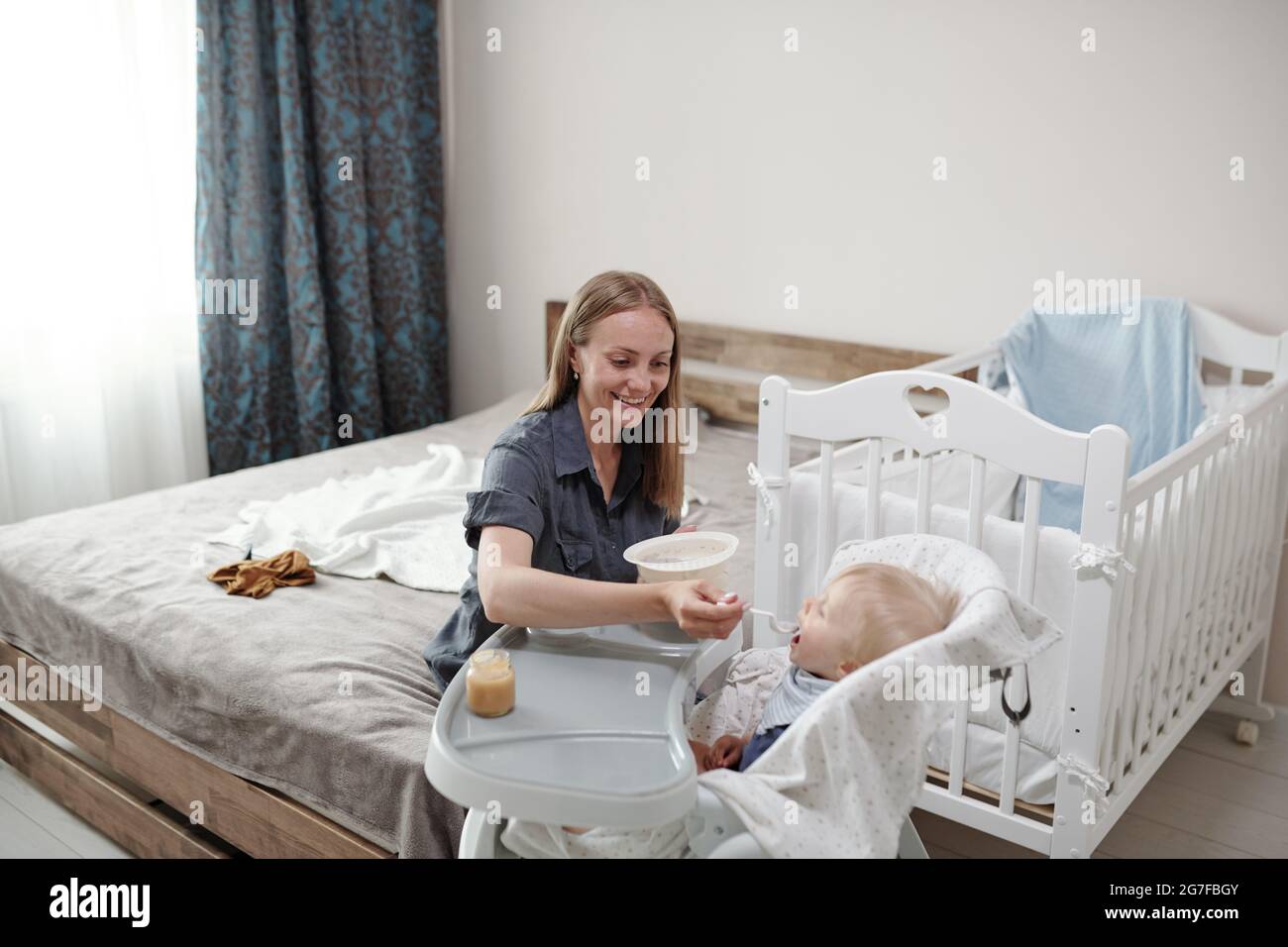 Glückliche junge Mutter im Mutterschaftsurlaub sitzt auf dem Bett und füttert das Baby im Hochstuhl, während sie sich um das Baby kümmert Stockfoto