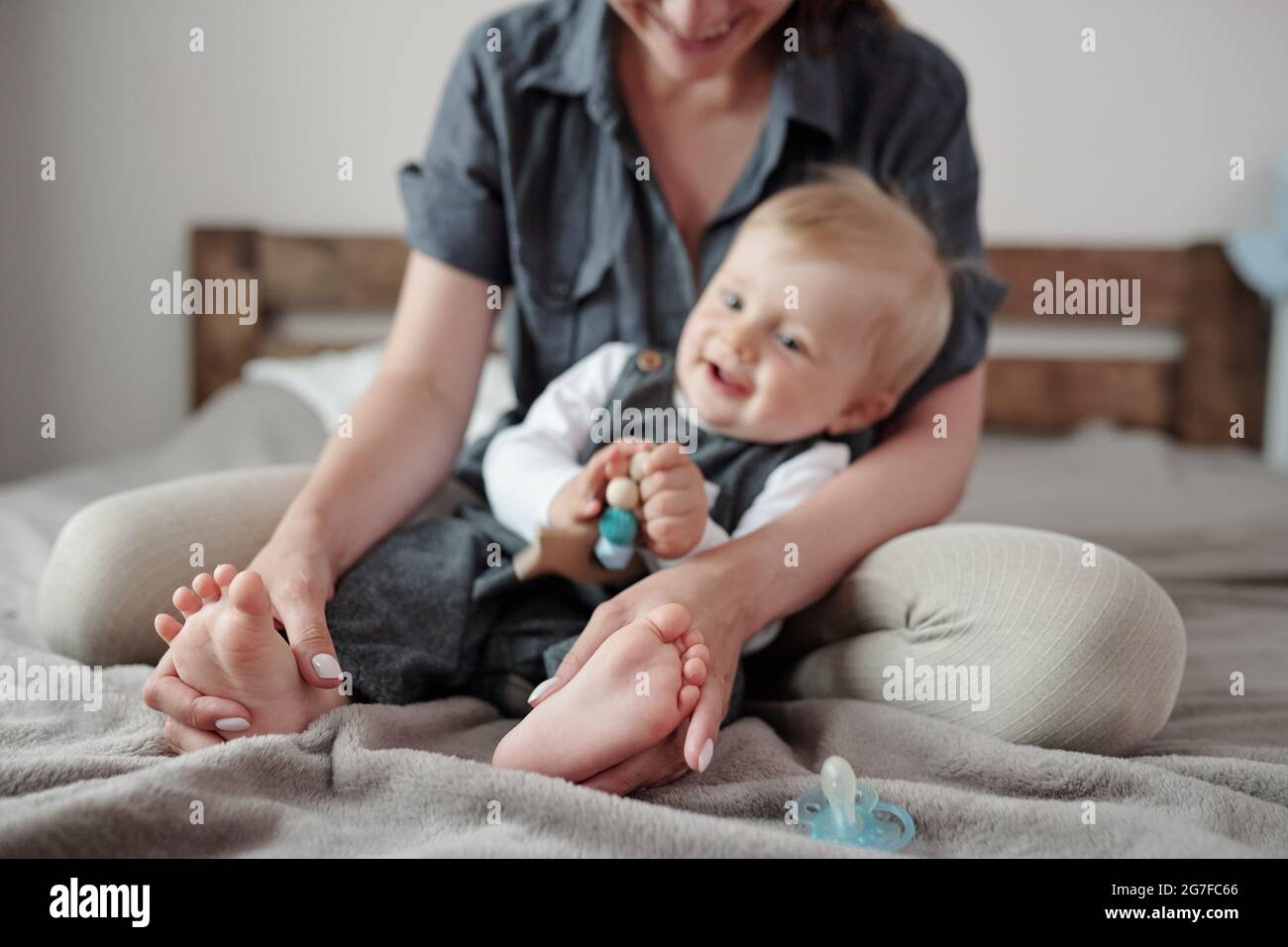 Lächelnde junge Mutter sitzt auf dem Bett und spielt mit Babys Füßen im Schlafzimmer Stockfoto