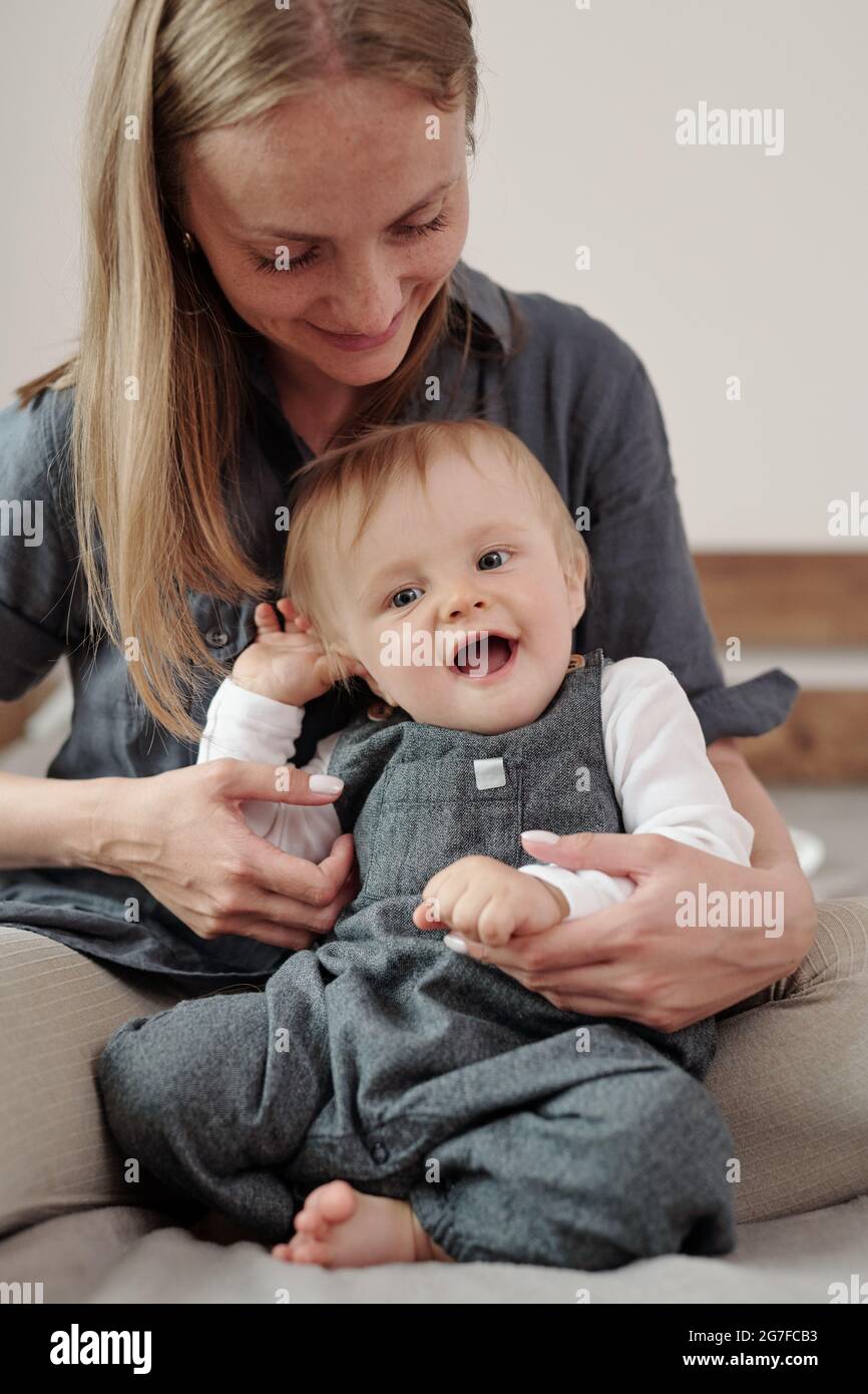 Lächelnde junge Mutter, die auf dem Bett sitzt und die Arme ihres Babys berührt Stockfoto
