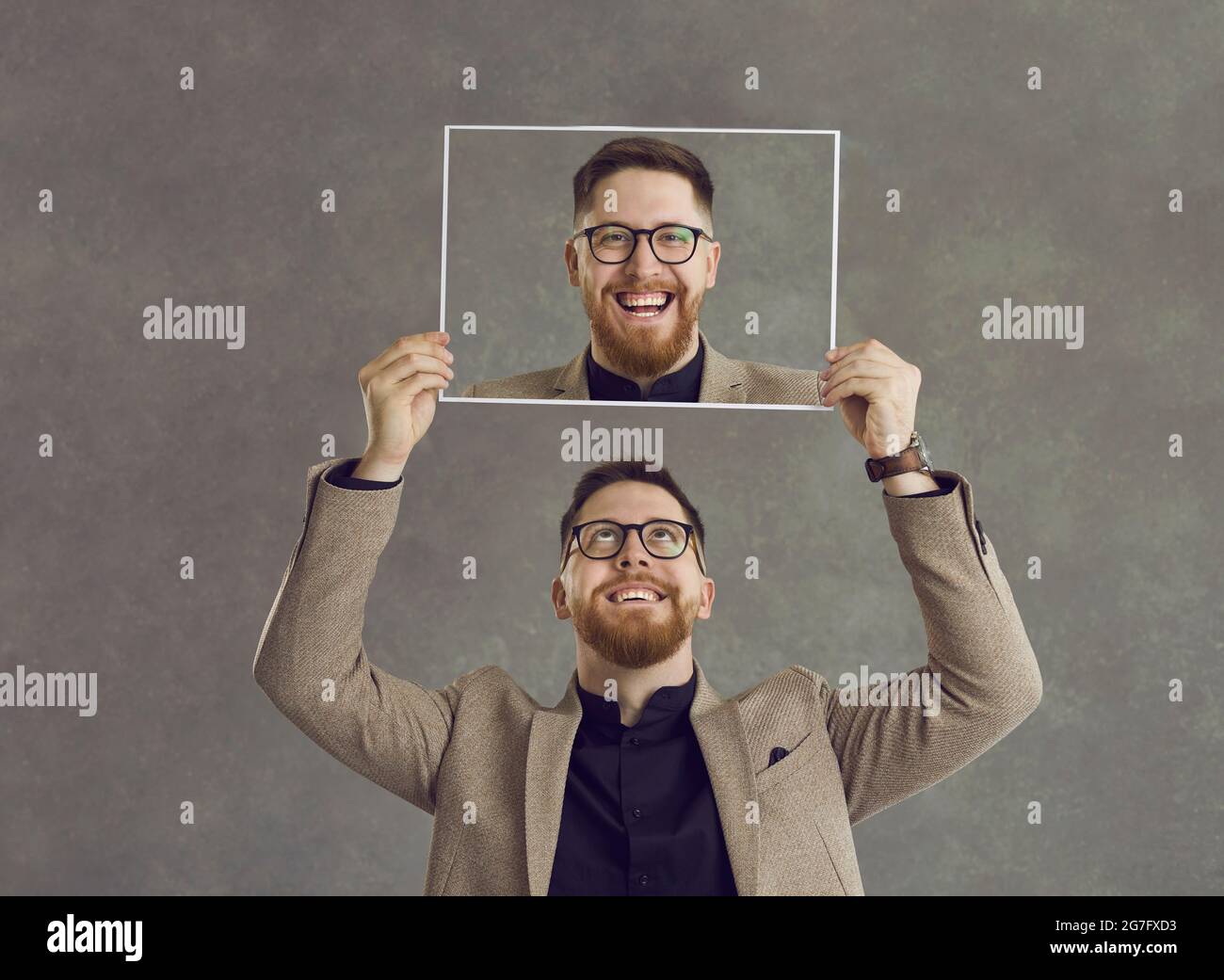 Glücklicher lächelnder Mann, der ein Foto von sich selbst hält, das isoliert auf einem grauen Hintergrund lacht Stockfoto Glücklicher lächelnder Mann, der ein Foto von sich selbst hält, das isoliert auf einem grauen Hintergrund lacht Stockfoto