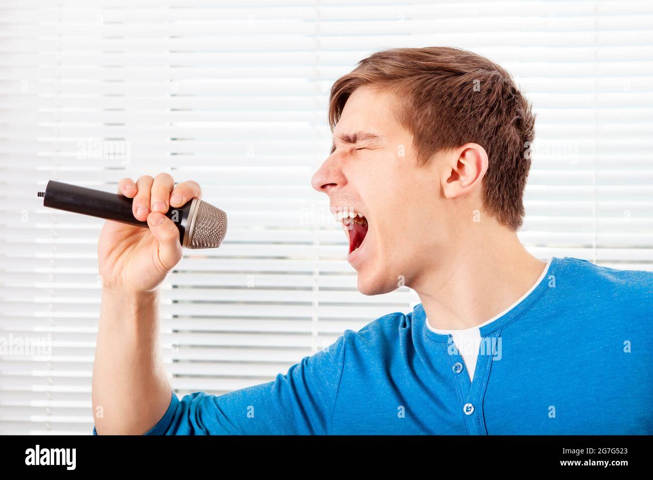 Junger Mann mit Mikrofon singt ein Lied auf dem Jalousie Hintergrund Stockfoto