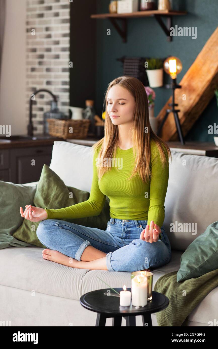 Barfuß sitzende Frau auf dem Sofa in Lotus-Pose und meditiert während der Yoga-Sitzung zu Hause Stockfoto