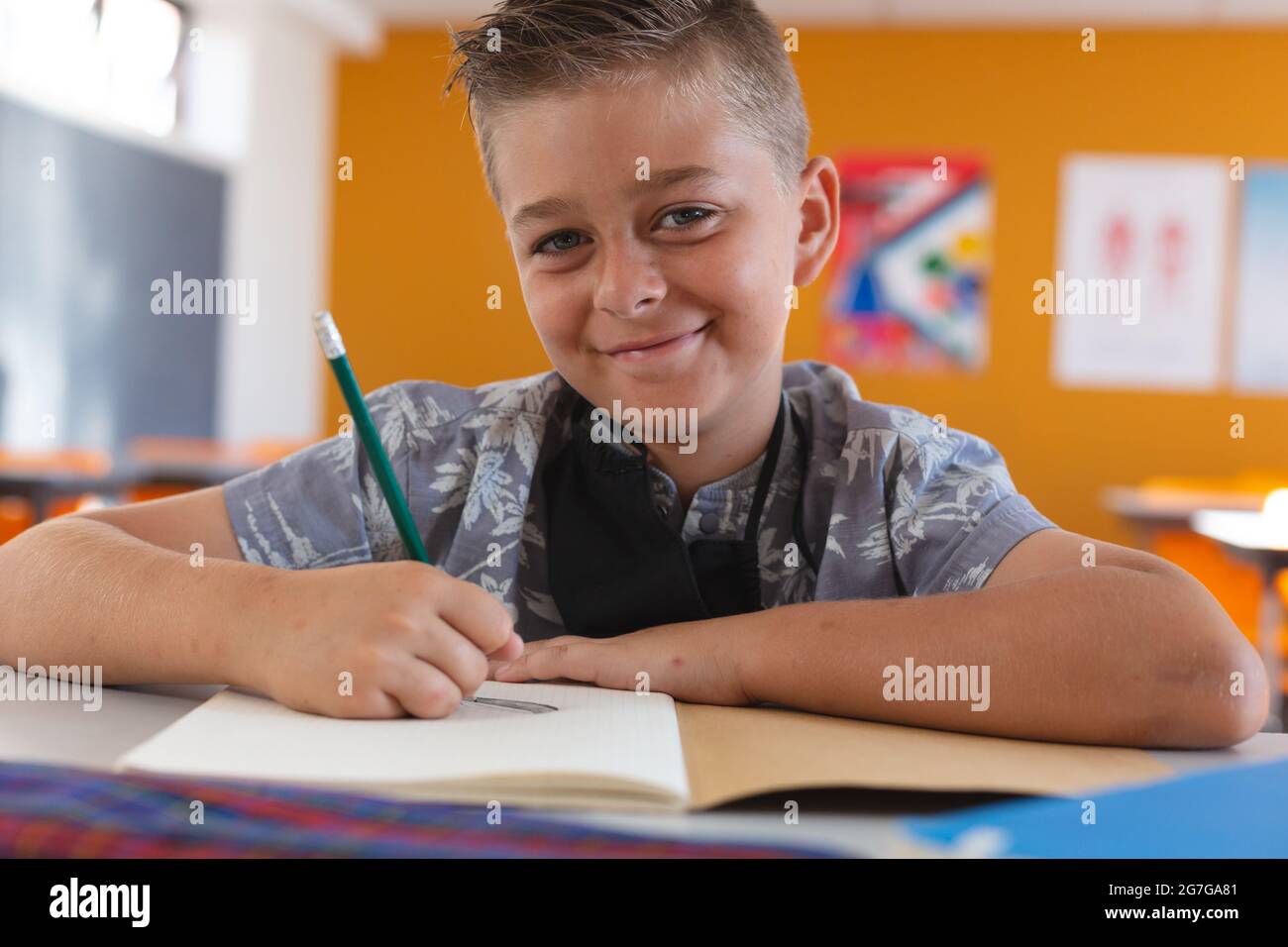Porträt eines lächelnden kaukasischen Schuljungen mit Gesichtsmaske, die im Klassenzimmer sitzt und in einem Schulbuch schreibt Stockfoto