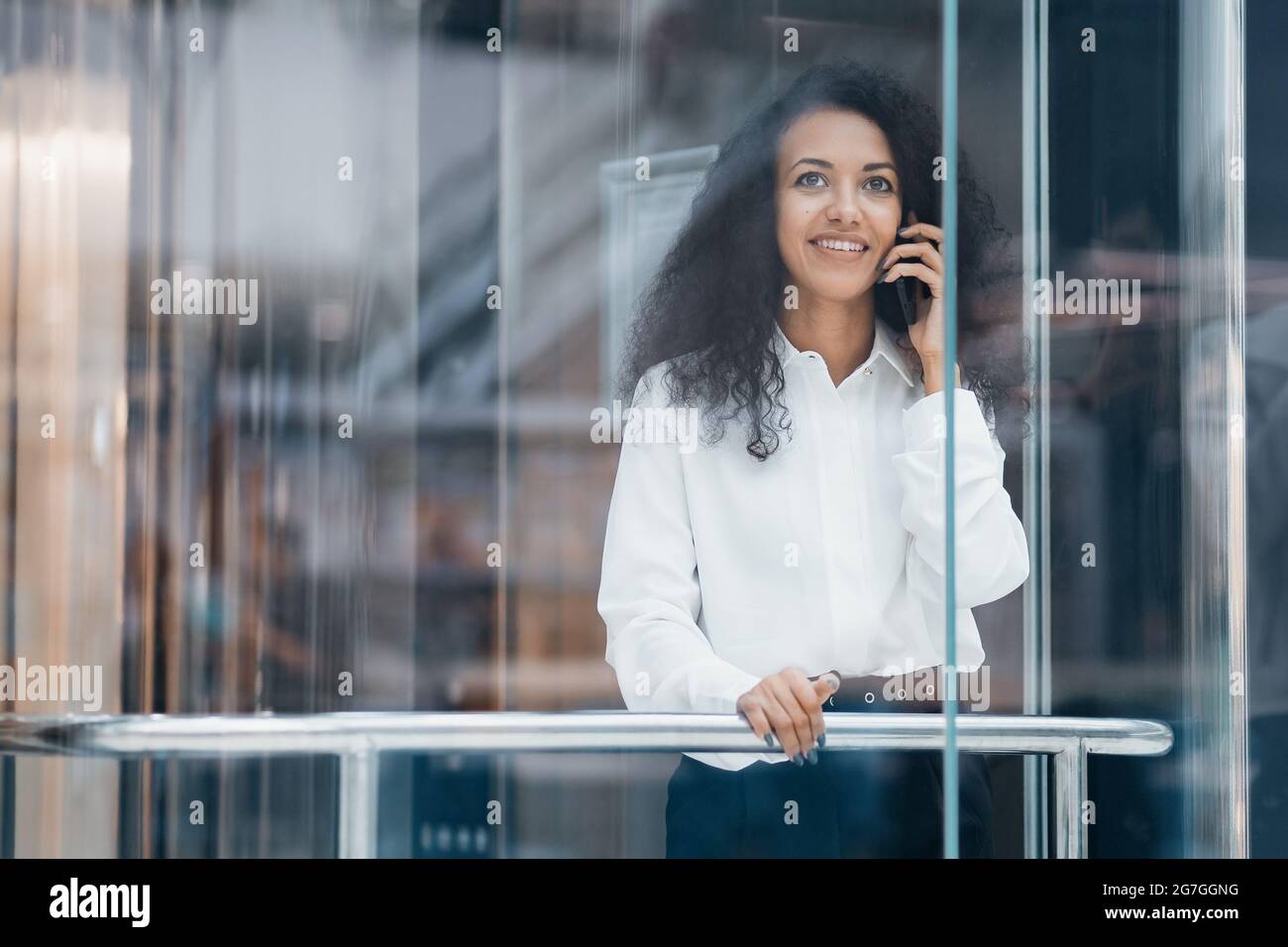 Durch das Glas. Junge Frau, die auf ihrem Smartphone spricht, während sie im Aufzug steht. Stockfoto