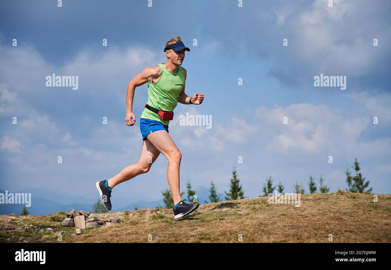 Voller muskulöser junger Mann, der den Hügel mit blauem bewölktem Himmel auf dem Hintergrund hochläuft. Schöner Läufer mit Sportshorts und Tanktop beim Joggen im Freien in den Bergen. Stockfoto