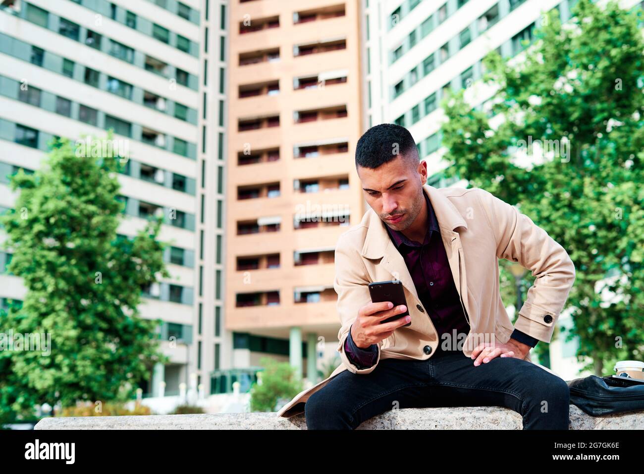 Ein niedriger Winkel von positiv gut gekleideten jungen hispanischen Geschäftsmann, der auf dem Smartphone SMS schreibt und Nachrichten auf der städtischen Straße mit zeitgenössischen Gebäuden diskutiert Stockfoto