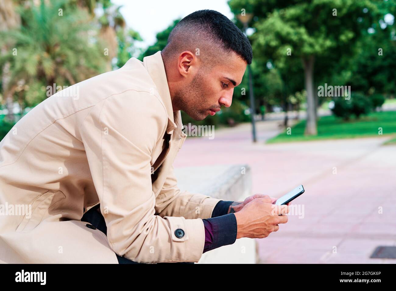 Ein niedriger Winkel von positiv gut gekleideten jungen hispanischen Geschäftsmann, der auf dem Smartphone SMS schreibt und Nachrichten auf der städtischen Straße mit zeitgenössischen Gebäuden diskutiert Stockfoto