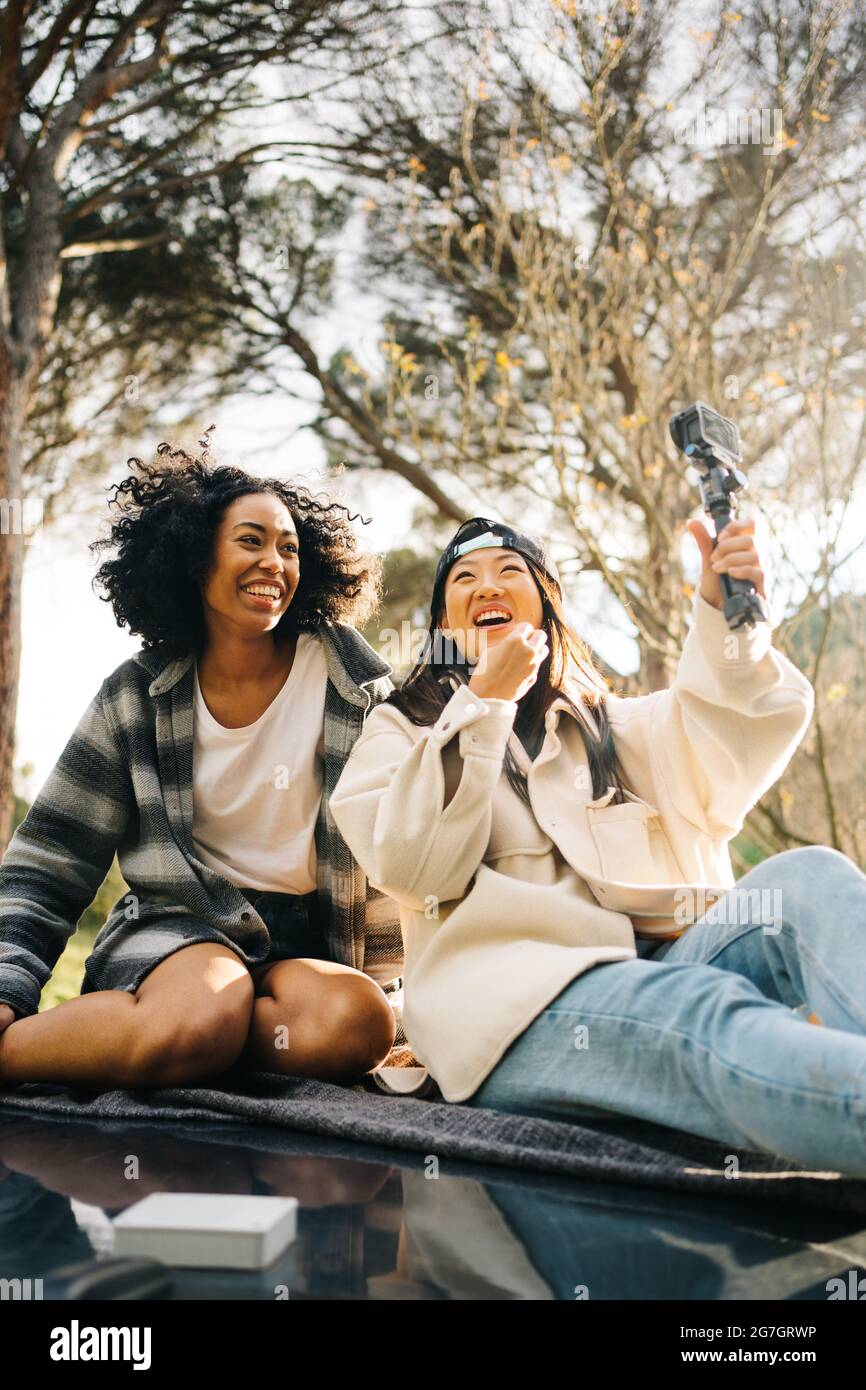 Ein niedriger Winkel von fröhlichen jungen multirassischen Freundinnen, die auf dem Dach eines Wohnwagens sitzen und Fotos machen, während sie gemeinsam den Sommerurlaub verbringen Stockfoto