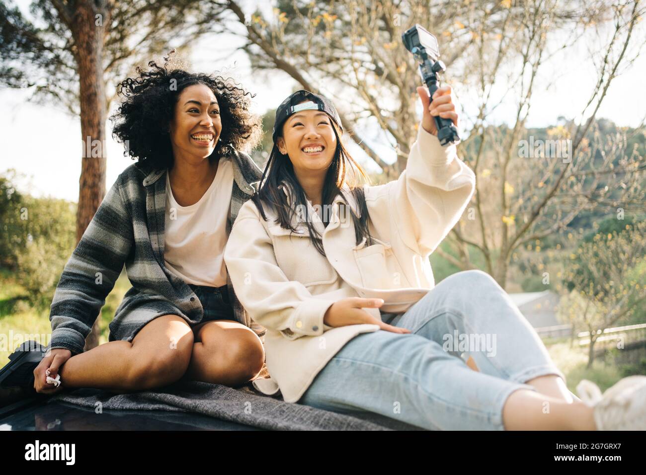 Ein niedriger Winkel von fröhlichen jungen multirassischen Freundinnen, die auf dem Dach eines Wohnwagens sitzen und Fotos machen, während sie gemeinsam den Sommerurlaub verbringen Stockfoto