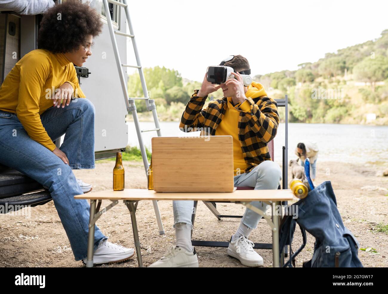Fröhlicher Mann in VR-Brille erkunden virtuelle Realität in der Nähe erfreut schwarze Freundin während in der Natur zusammen ruhen Stockfoto