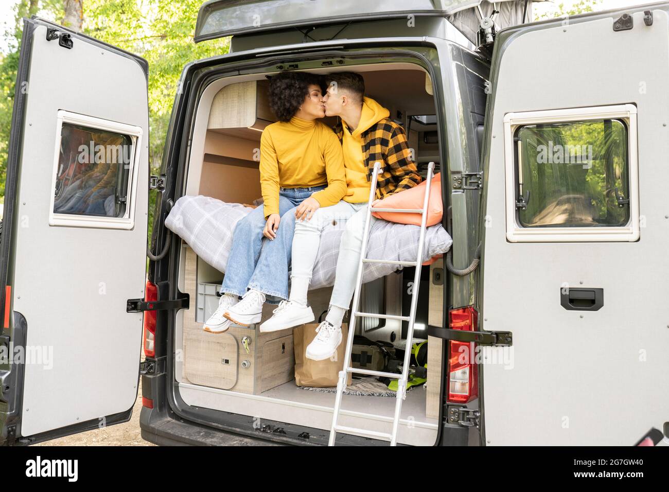 Verschiedene Männer und Frauen küssen sich gegenseitig, während sie auf dem Bett in einem modernen Van auf der Straße sitzen Stockfoto