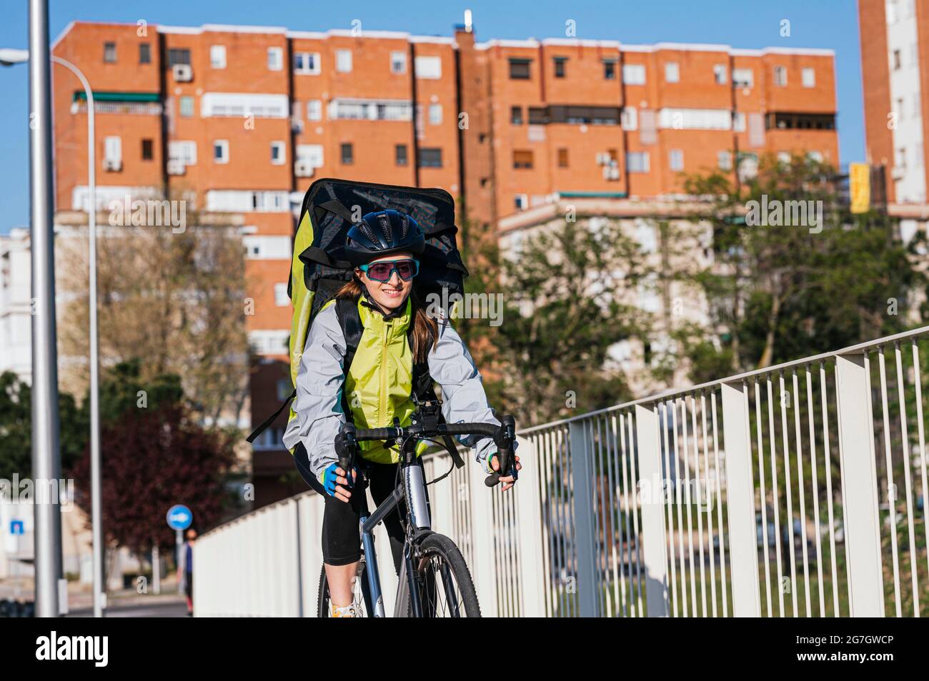 Fröhliche Kurierin mit Thermotasche, die Fahrrad auf der Brücke fährt, während sie Essen in der Stadt liefert Stockfoto