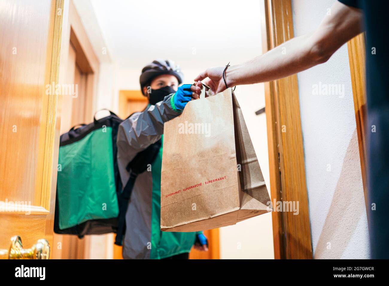 Delivery Frau mit thermischen Rucksack und Gesichtsmaske geben Papiertüte mit Lebensmitteln gesichtslosen Kunden durch Wohnungstür zu ernten Stockfoto