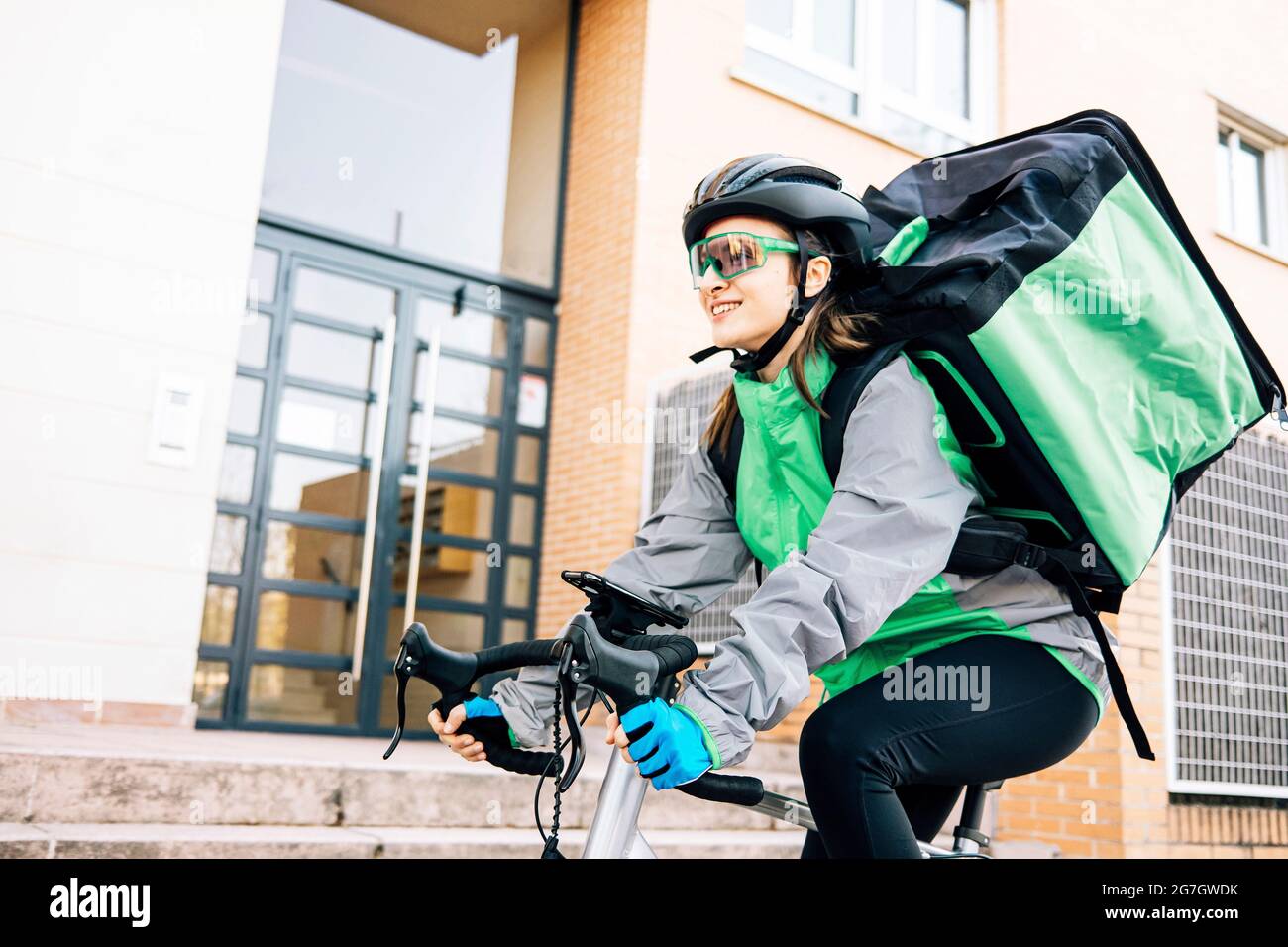 Niedriger Winkel der weiblichen Kurierin mit Thermobeutel lächeln und Fahrrad fahren auf der Straße, während die Lieferung an sonnigen Tag in der Stadt Stockfoto
