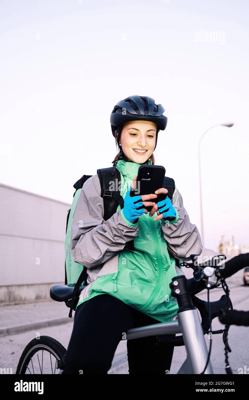 Junge, fröhliche Frau mit Thermotasche, die auf dem Smartphone mit GPS-Karte unterwegs ist, während sie auf dem Fahrrad sitzt, um Essen auf der Straße der Stadt zu liefern Stockfoto