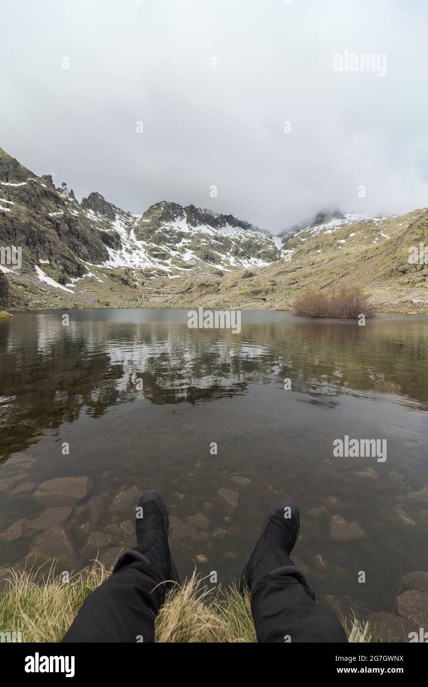 POV des anonymen Touristen, der sich an der Küste des friedlichen Sees Laguna Grande im circo de Gredos, umgeben von verschneiten Bergen in Avila, Spanien, ausruhte Stockfoto