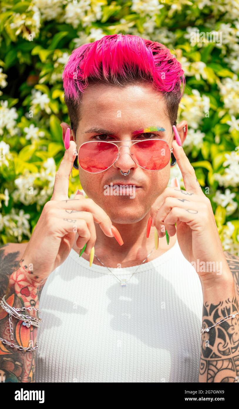 Entschlossener schwuler Mann mit gefärbtem rosa Haar und langen bunten Nägeln, die Sonnenbrille berühren und die Kamera im Sommerpark betrachten Stockfoto