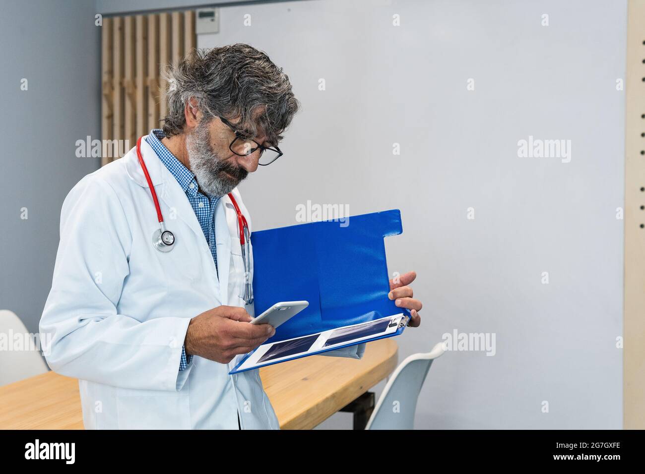 Fokussierter älterer männlicher Sanitäter in Uniform und mit Röntgenstrahlen in einem Ordner, der im Krankenhaus steht und auf dem Mobiltelefon surft Stockfoto