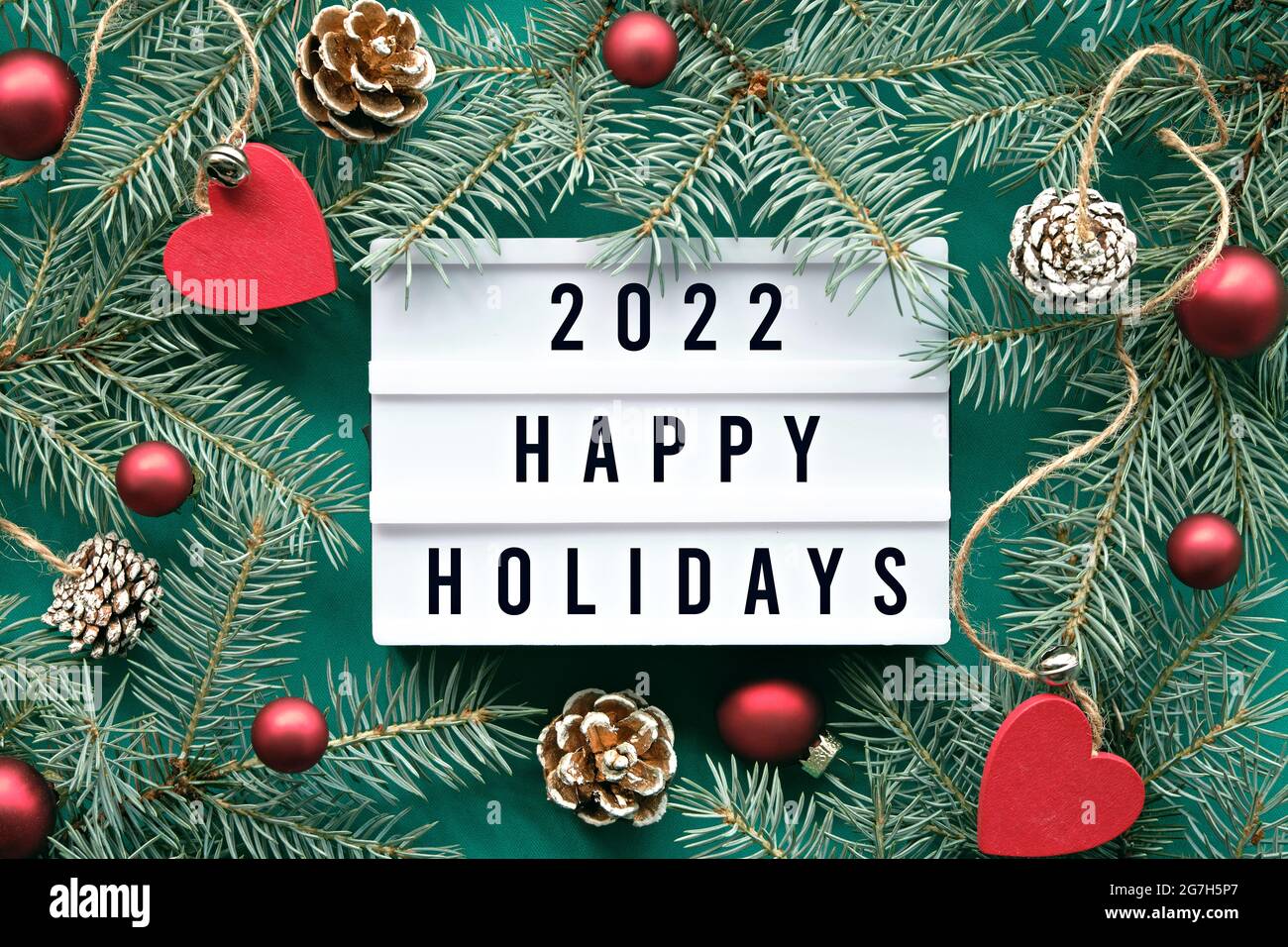 Leuchttafel mit Text 2022 Happy Holidays. Flach legen mit Tannenzweigen und Weihnachtsdekorationen um Leuchtkasten. Tannenzapfen, rote Holzherzen und Glas Stockfoto