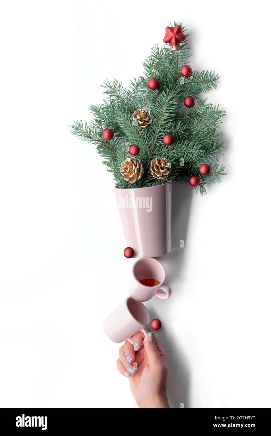 Kreative Xmas Balance Arrangement. Kaffeetassen unterstützen Blumentopf mit Weihnachtsbaumzweigen, die mit roten Weihnachtskugeln und Tannenzapfen verziert sind. Handstützen Stockfoto