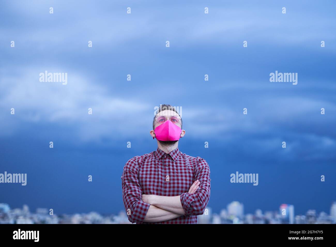 Konzept der Hoffnung, LGBTQ-Thema. Attraktive kaukasische Homosexuell in rosa schützende Gesichtsmaske und rosa Brillen Blick nach oben mit Hoffnung. Männliche Person Porträt mit regnerischem Wetter Hintergrund und städtische Skyline Stockfoto