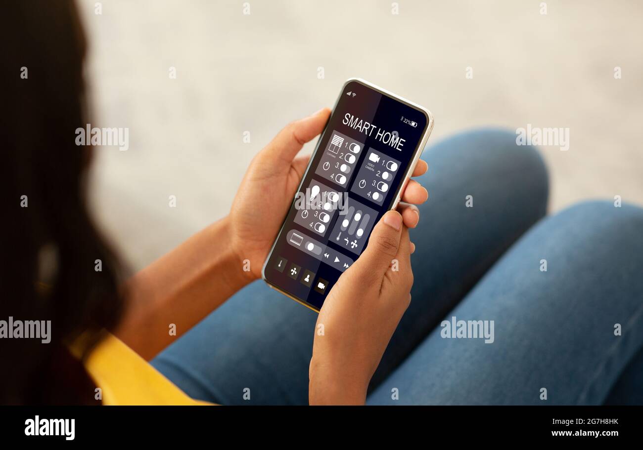 Frau steuert App mit Smartphone, Nahaufnahme auf mobilen Gerät mit gestartet Smart Home. afroamerikanische Weibchen konfiguriert Hausfunktionen für comf Stockfoto