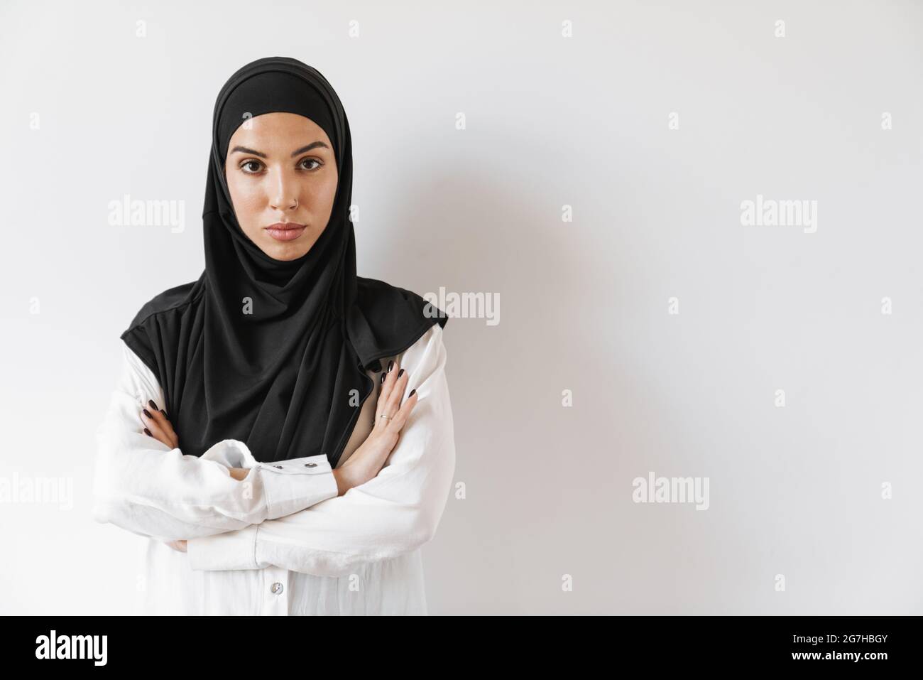 Eine selbstbewusste muslimische Frau mit durchbohrter Nase, die mit gekreuzten Armen im weißen Studio steht Stockfoto