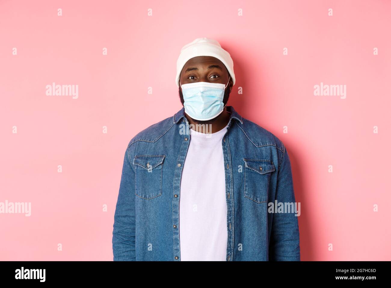 Covid-19, Lifestyle und Quarantänekonzept. Happy Black Kerl in Mütze und Gesichtsmaske lächelnd mit Augen, stehen über rosa Hintergrund Stockfoto
