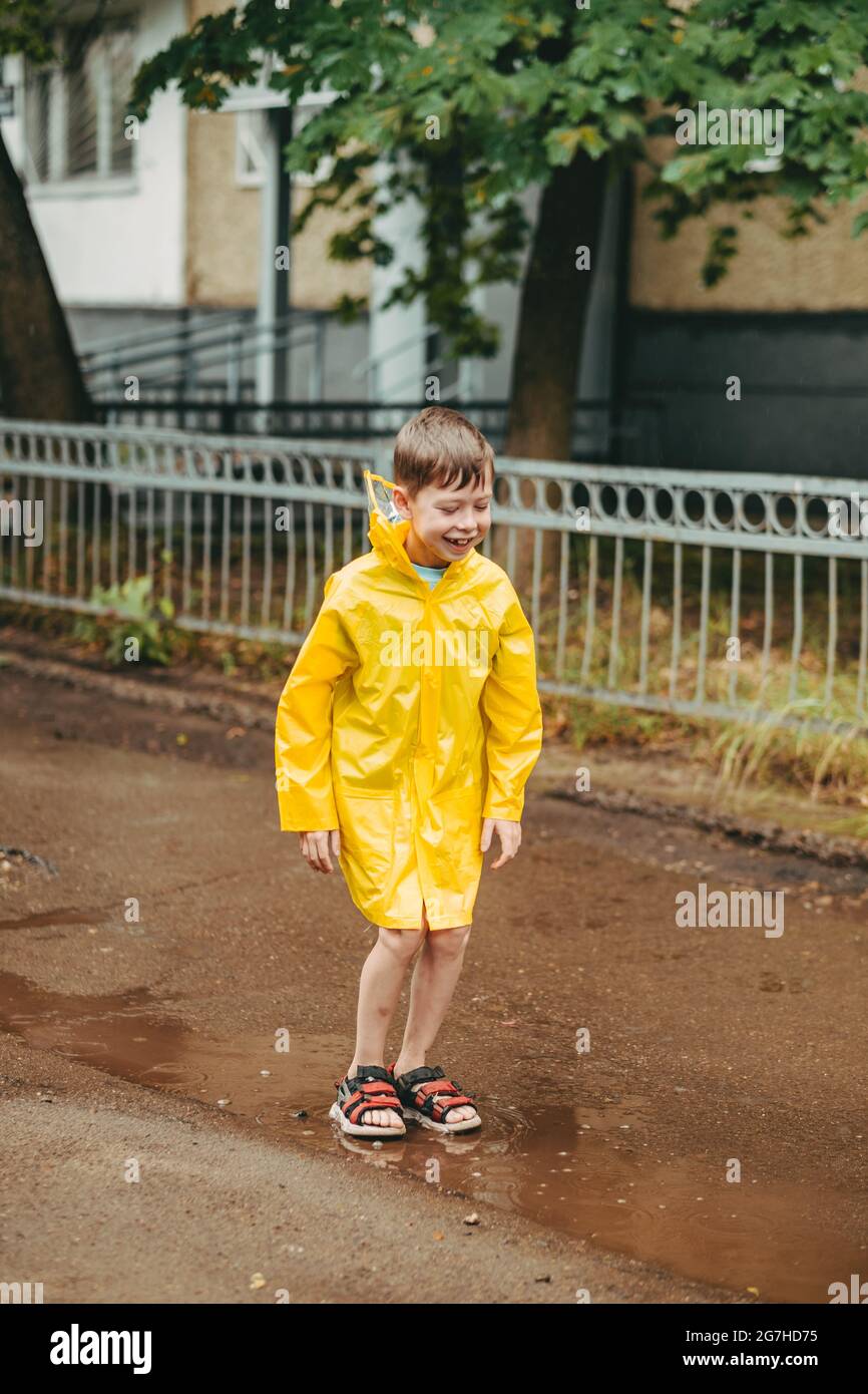 Ein Junge in einem gelben Regenmantel geht im Regen draußen. Ein Kind springt auf die engen Wege. Springt durch schlammige Pfützen. Glückliche und unbeschwerte Kindheit Stockfoto