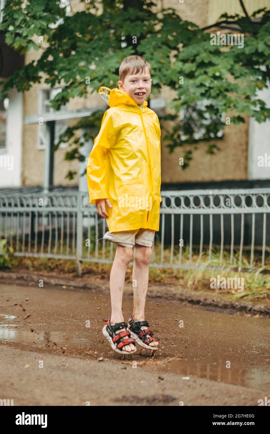 Ein Junge in einem gelben Regenmantel geht im Regen draußen. Ein Kind springt auf die engen Wege. Springt durch schlammige Pfützen. Glückliche und unbeschwerte Kindheit Stockfoto