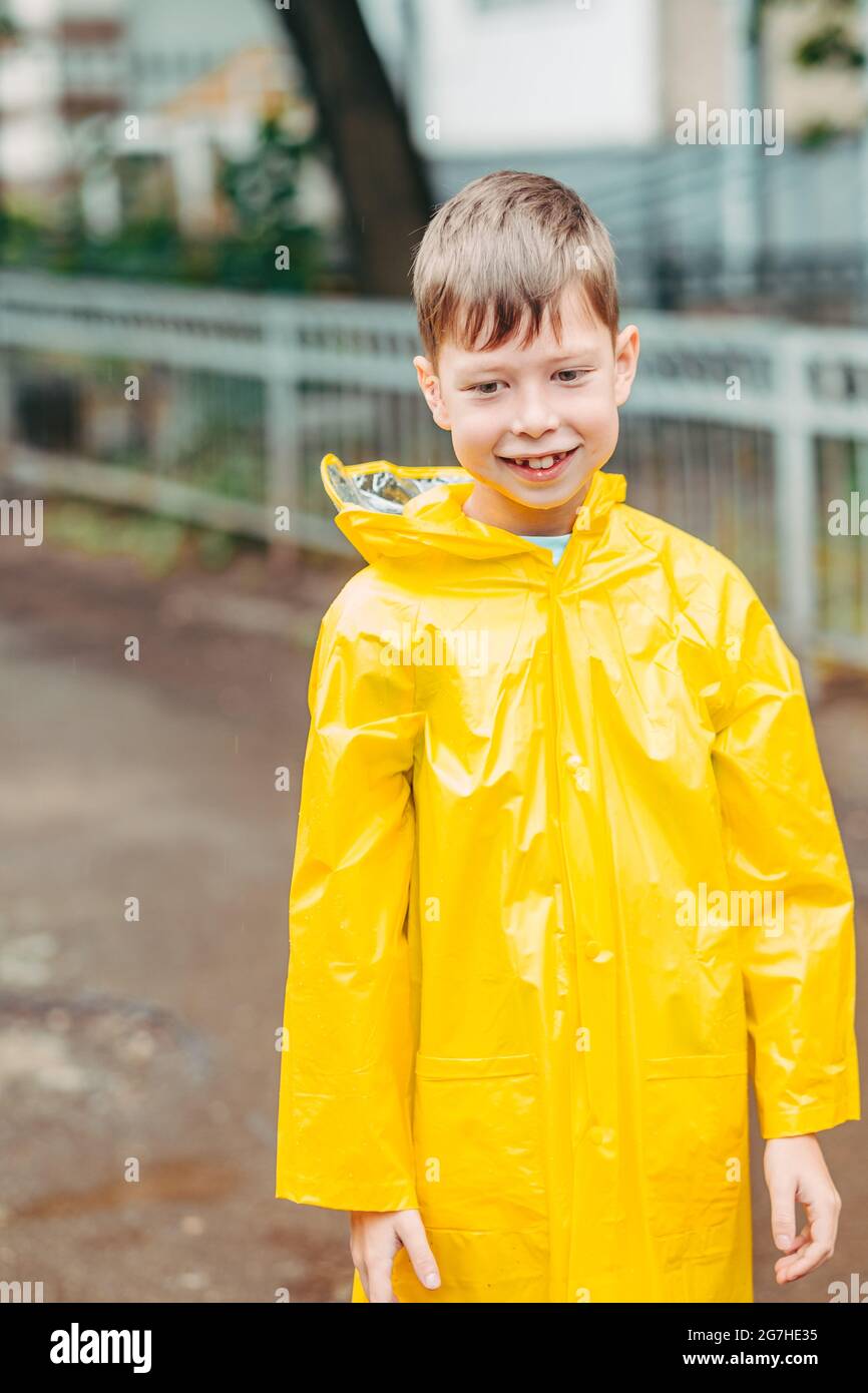 Ein Junge in einem gelben Regenmantel geht im Regen draußen. Schönes und lächelndes Kind auf der Straße. Porträt eines Jungen von europäischer Unbehaglichkeit. Öffnen Stockfoto