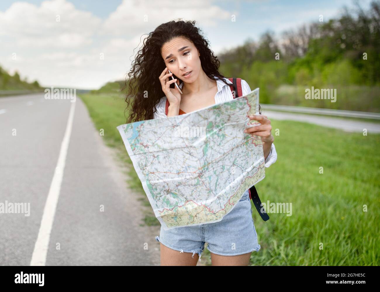 Gestresste weibliche Touristin mit Karte, die am Straßenrand steht, auf dem Smartphone spricht, kann weder ein Auto erwischen noch ihre Reiseroute finden. Junge Dame fühlt sich verloren whil Stockfoto