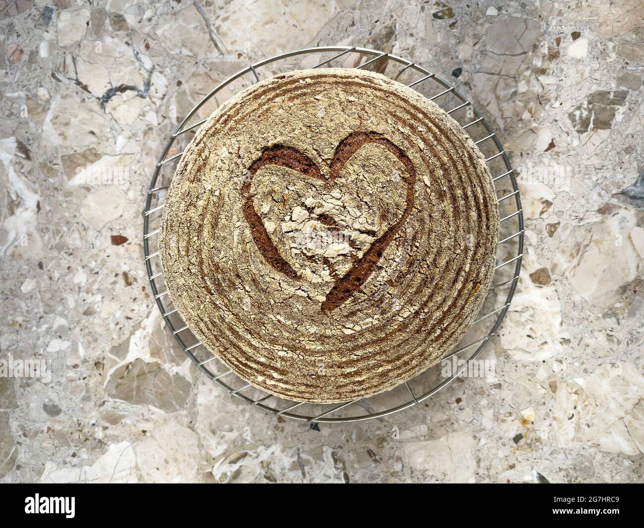 Grundansicht eines runden Sauerteig-Laibs Brot, das in der Kruste herzförmig geritzt ist und auf einem Drahtgestell auf einer Marmorarbeitsfläche abkühlt. Stockfoto