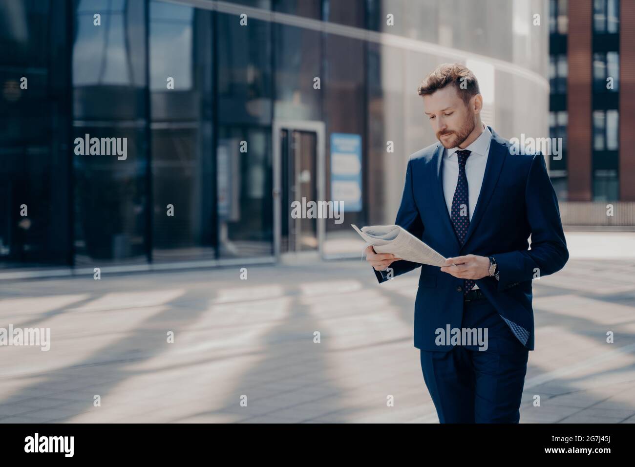 Fokussierter, formell gekleideter Geschäftsmann, der Zeitung liest, während er in der Nähe des Bürogebäudes läuft Stockfoto