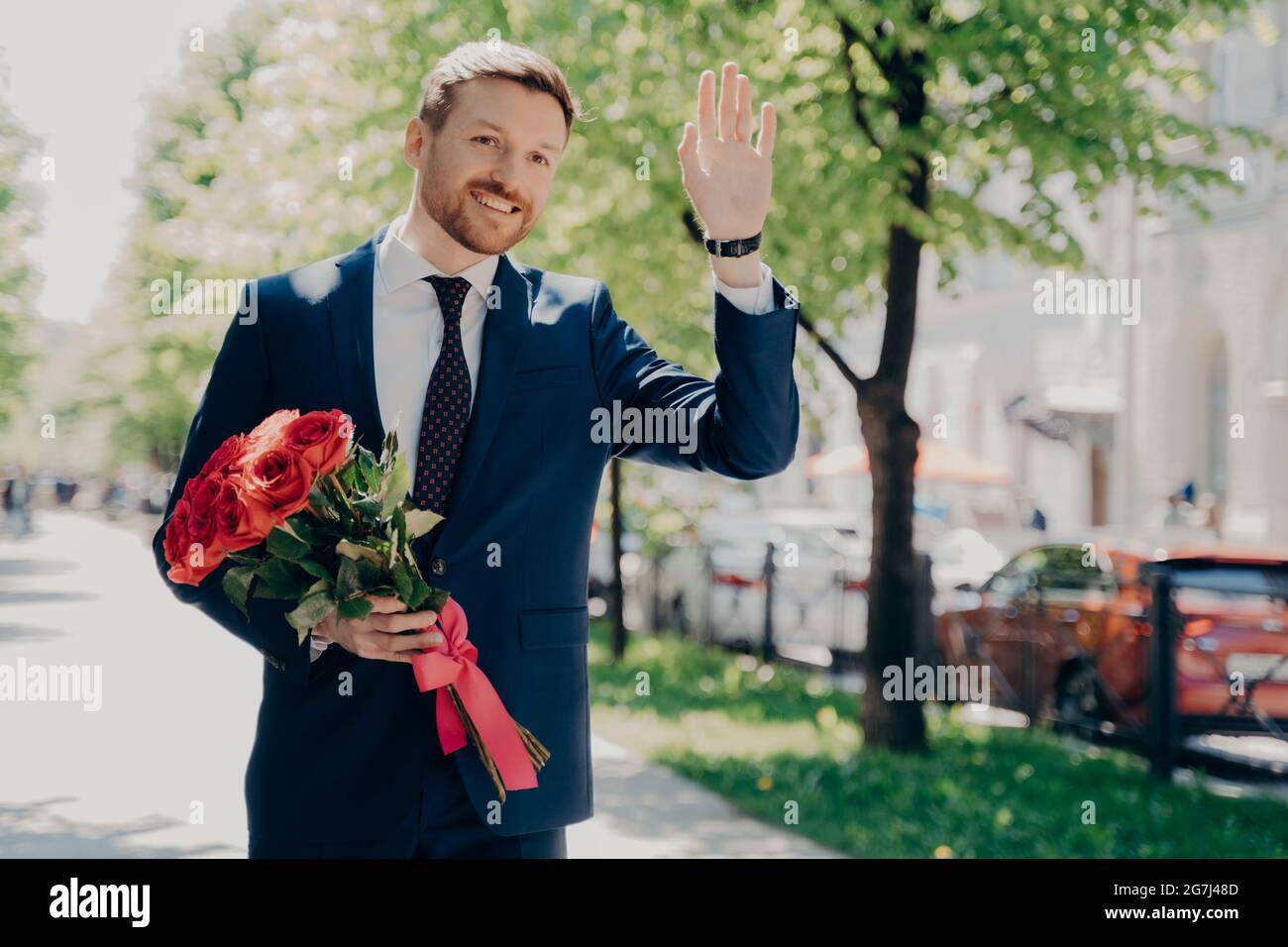 Fröhlicher, männlicher Geschäftsmann mit Blumenstrauß, der jemanden im Stadtpark begrüßt Stockfoto