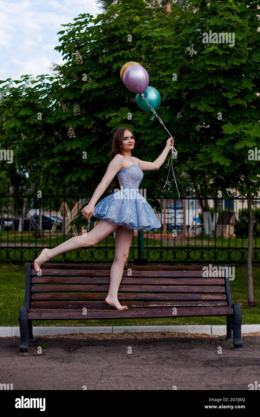 Eine lächelnde, langbeinige junge Frau in einem blauen kurzen Korsett-Kleid geht barfuß auf einer Bank und hält fliegende Ballons Stockfoto