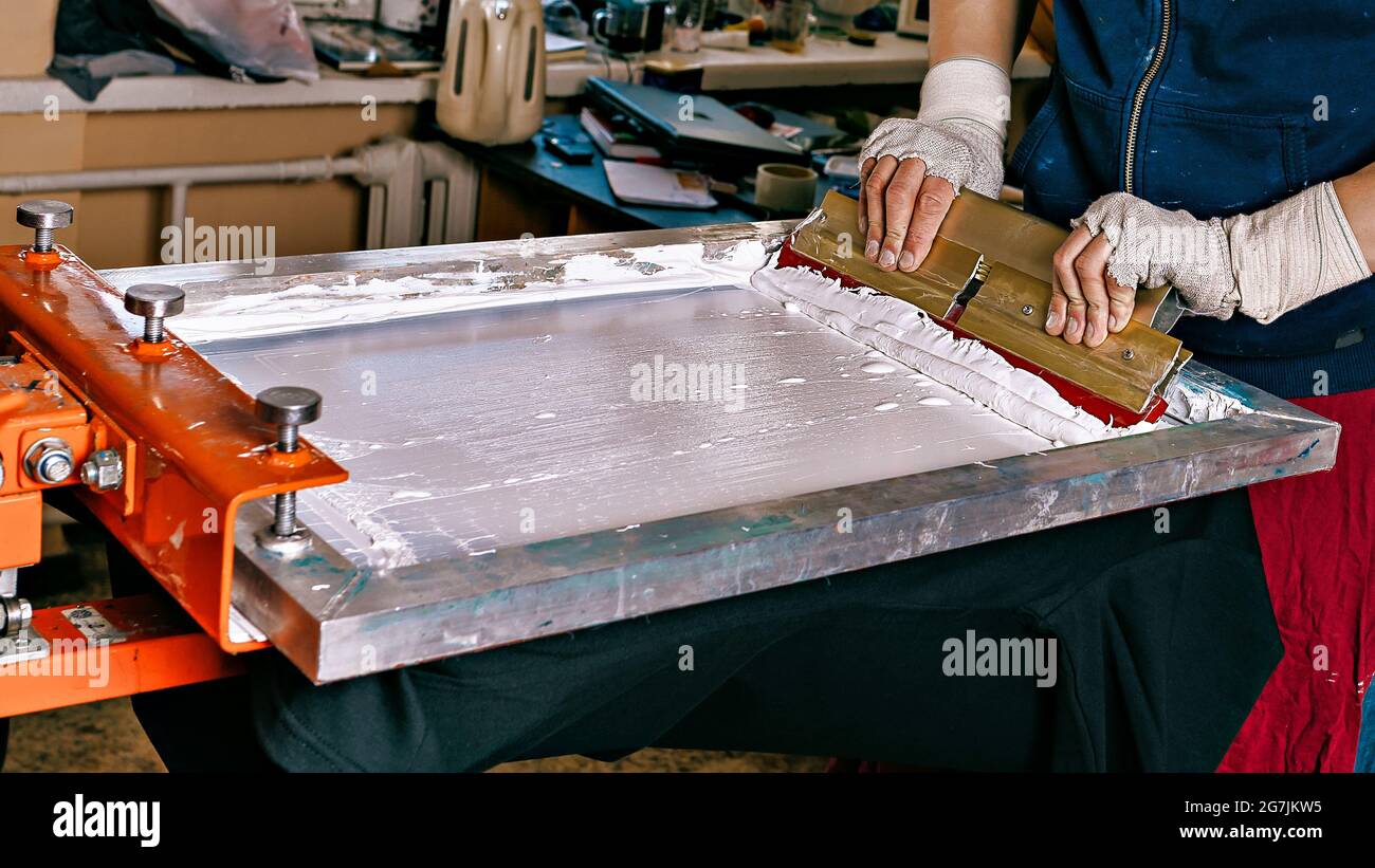 Selektives Fokusfoto von männlichen Händen mit einem Rakel. Siebdruck-Produktion. Druck von Bildern auf Kleidung im Siebdruck-Verfahren in einem Designstudio Stockfoto