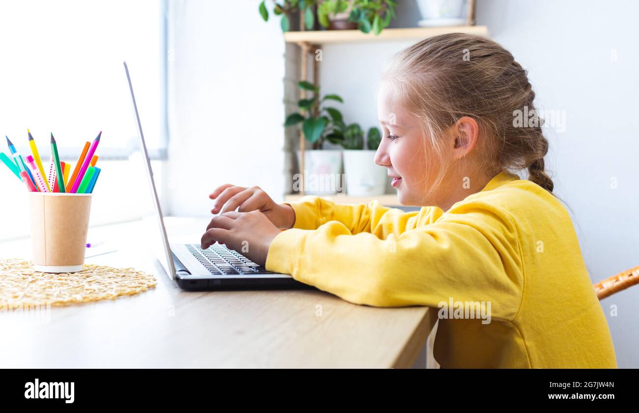 Kaukasisch blonde Schulmädchen, tippen auf einem Laptop-Tastatur, während Sie am Fenster zu Hause sitzen, Bloggen, Chatten, Anzeigen von Social-Media-Seiten oder Stockfoto