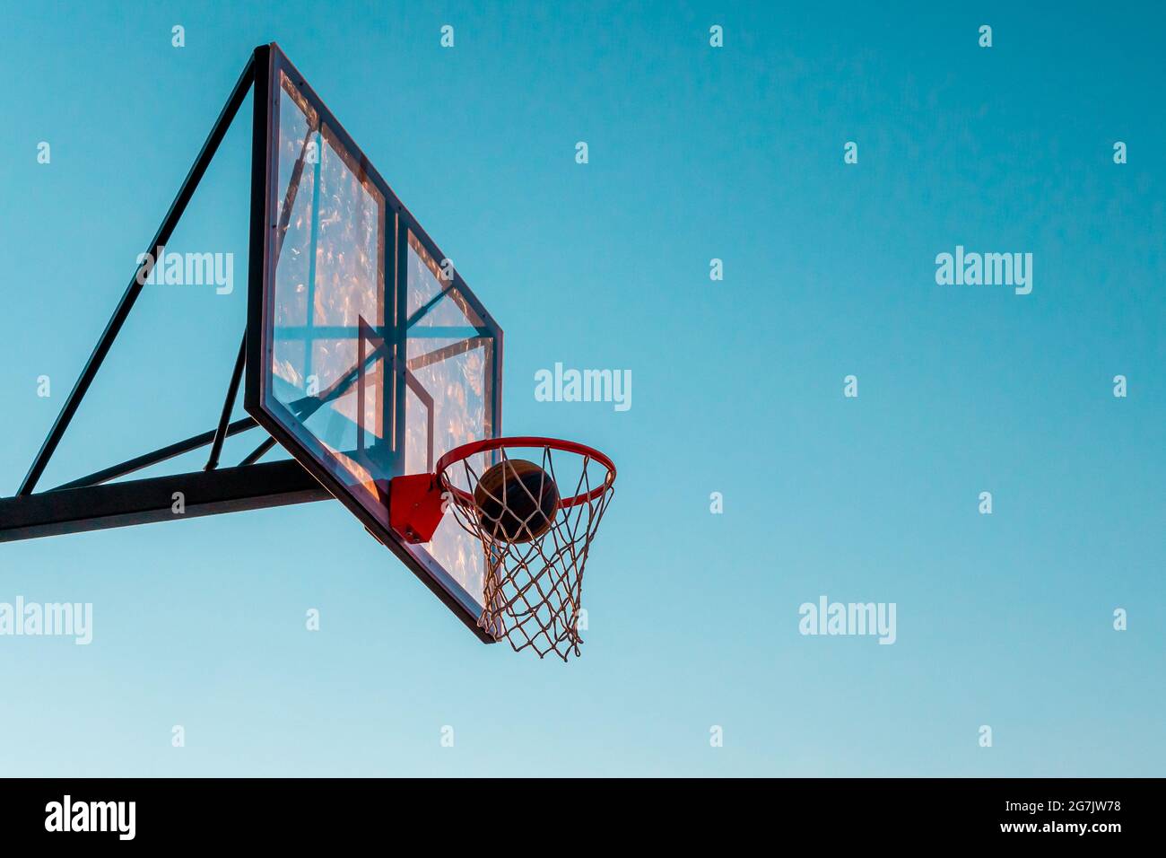 Ein Basketballkorb mit einem Ball auf einem blauen Himmel Hintergrund. Transparentes Kunststoff-Basketballschild auf dem Outdoor-Basketballfeld. Stockfoto